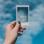 A hand with painted nails holds a Polaroid picture of clouds against a vibrant blue sky background.