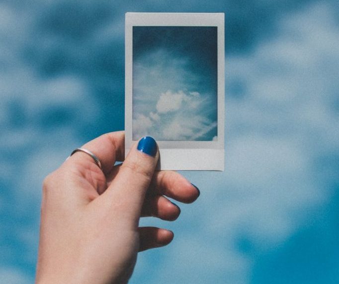 A hand with painted nails holds a Polaroid picture of clouds against a vibrant blue sky background.